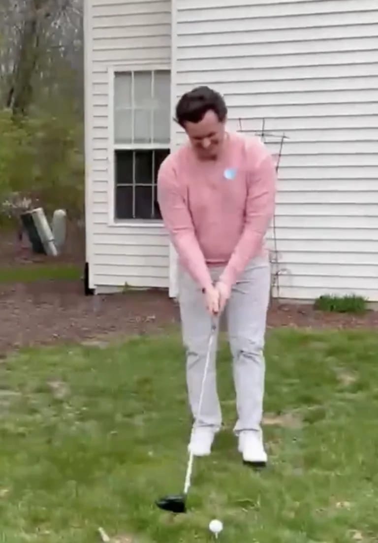 Tom Dillon addressing a gender reveal golf ball on a tee in a backyard, mid-swing setup