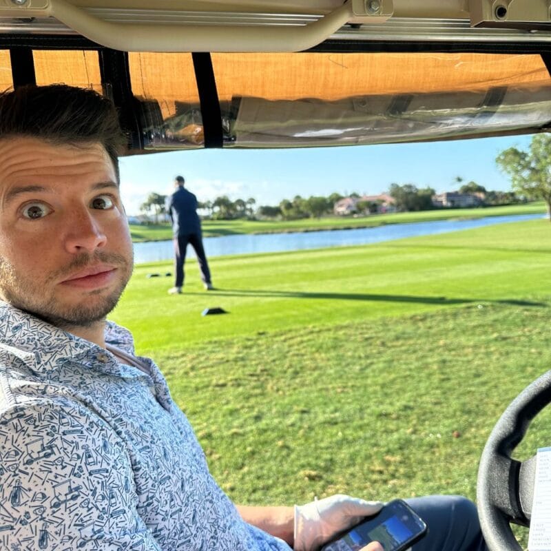 Tom Dillon sitting in a golf cart with a wide-eyed expression, golfer preparing to swing in the background on a sunny course
