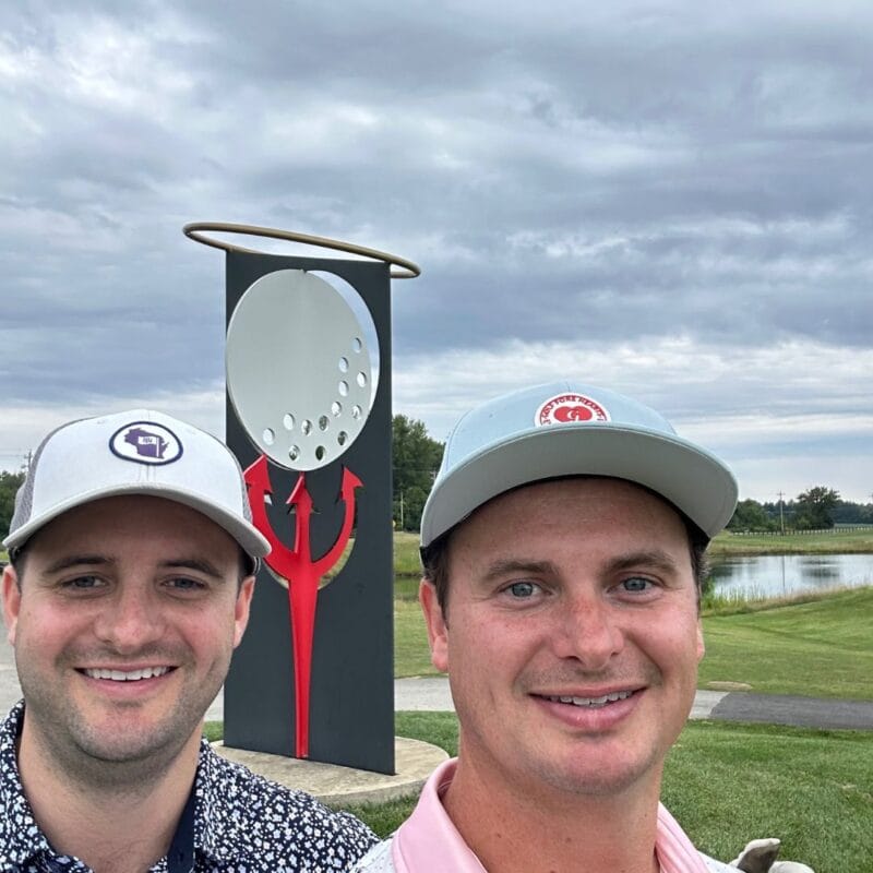 Two golfers posing in front of a large devil-and-golf-ball sculpture at a golf course under a cloudy sky