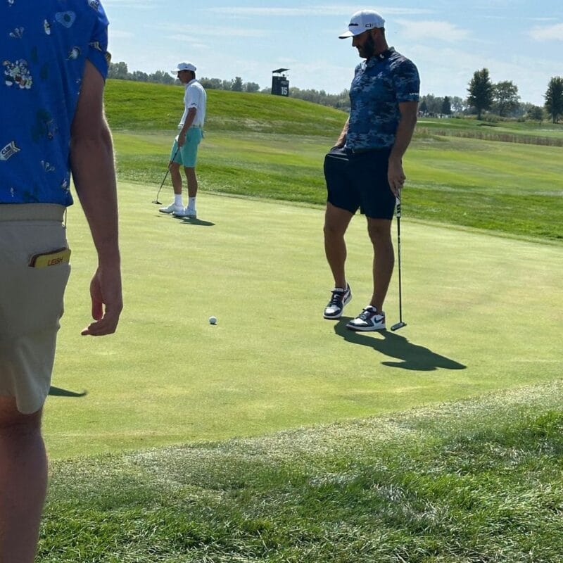 Three golfers on a sunny putting green, one lining up a putt while two others watch nearby