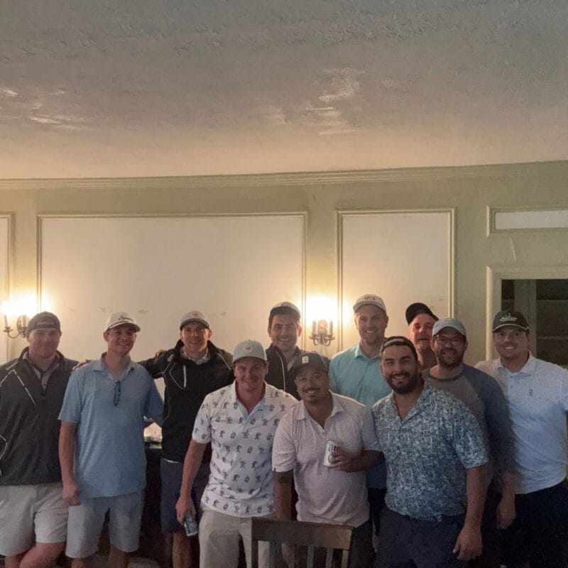 Group of about a dozen men in golf caps gathered inside a clubhouse for a group photo after a round