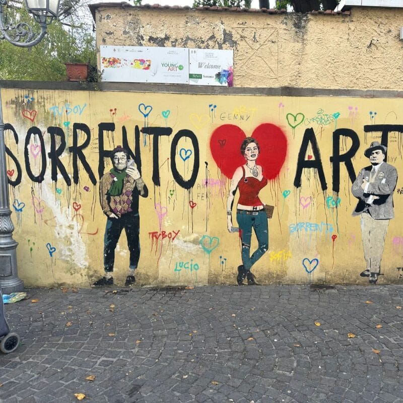 Street mural in Sorrento reading Sorrento Heart Art with painted figures on a yellow wall along a cobblestone street