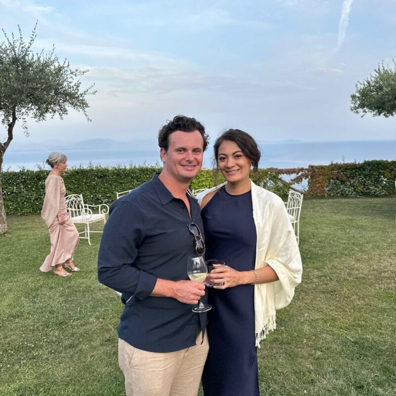 Tom Dillon and his partner holding wine glasses at an outdoor garden party on the Amalfi Coast, sea visible in the background