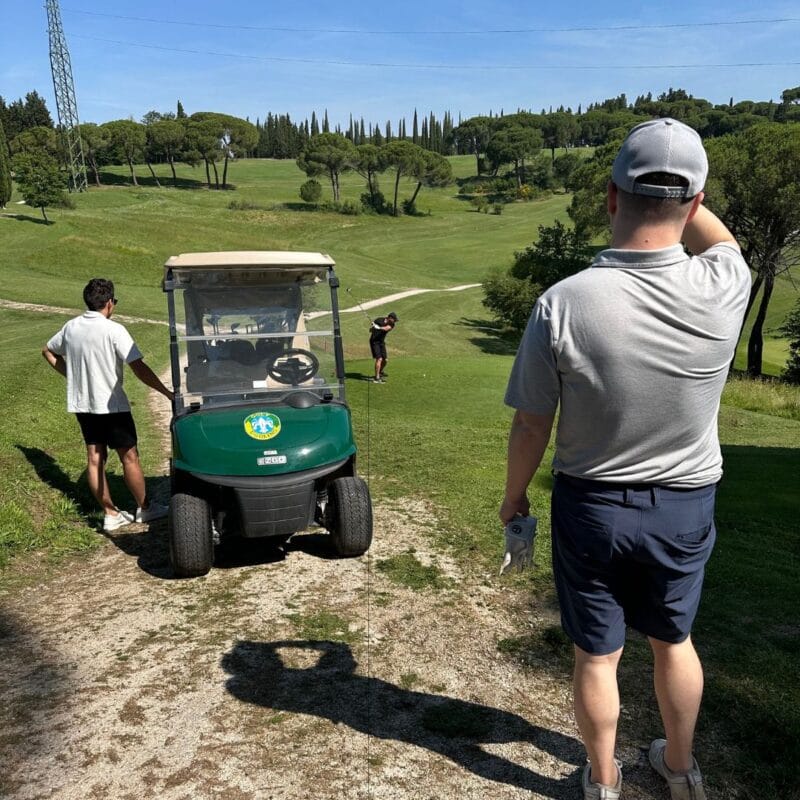 Two golfers watching a third tee off on an Italian golf course, with a green golf cart parked nearby