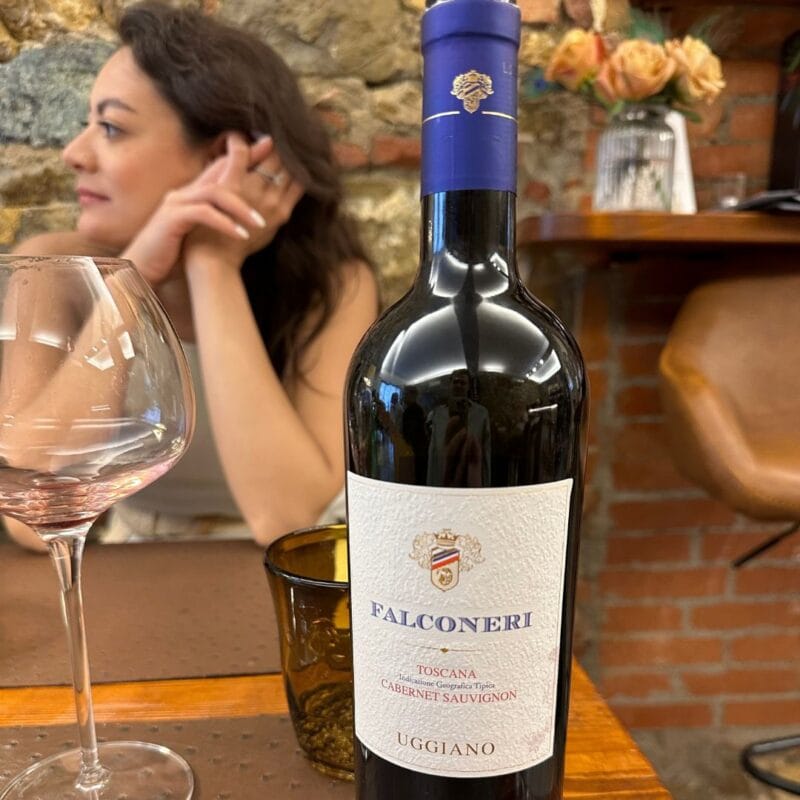 Bottle of Falconeri Toscana Cabernet Sauvignon by Uggiano on a wooden table at a Tuscan winery, woman seated in background